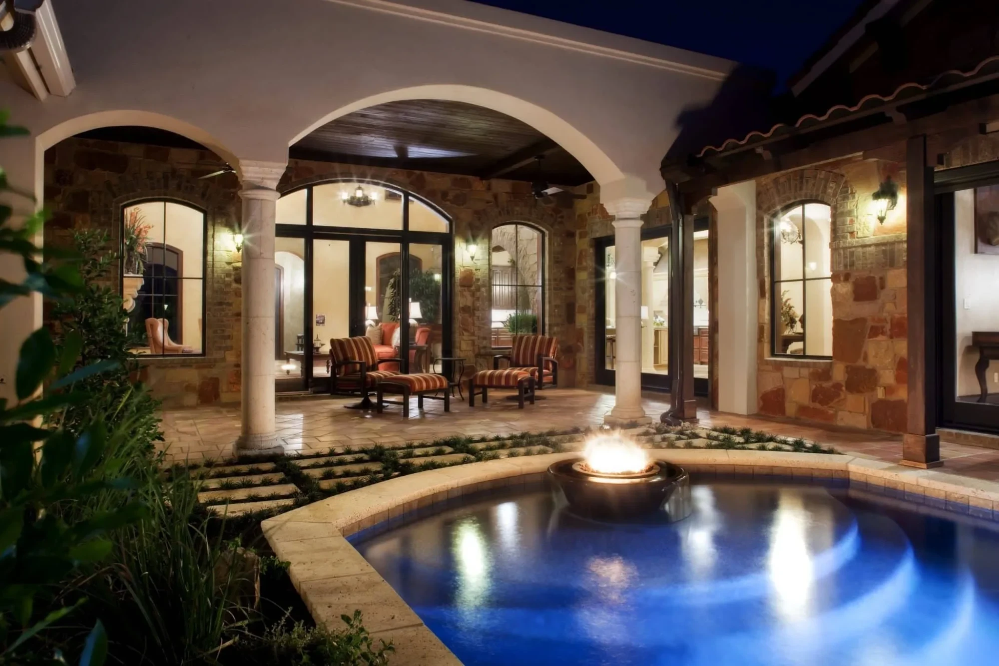 Courtyard pool with fire bowl at night beside an arched stone loggia, Mediterranean Parade Home