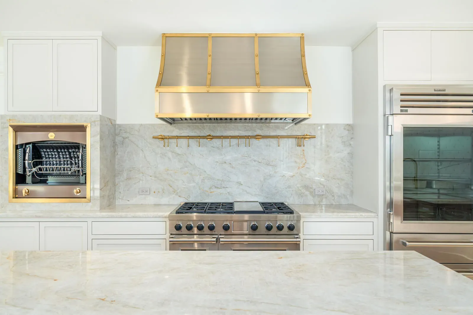 White show kitchen with brass range hood, marble backsplash, and professional gas range
