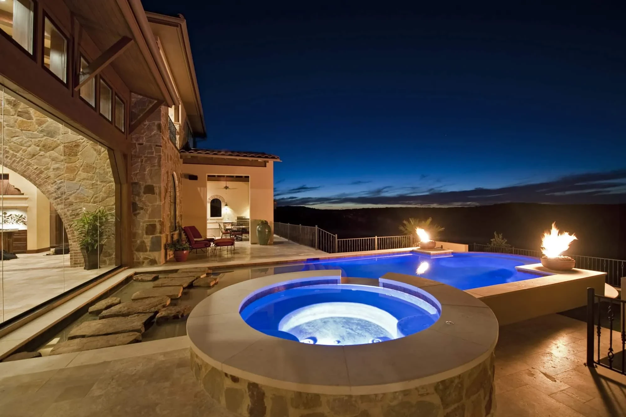 Round spa and infinity pool at night with fire bowls and dark hills beyond, Modern Mediterranean Parade Home